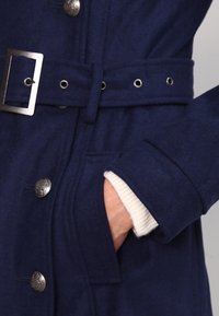 Mörkblå ullkappa med bälte i midjan, metallknappsstängning och fyra silverfärgade knappar. Har en ficka vid midjan och texturerad tyg.