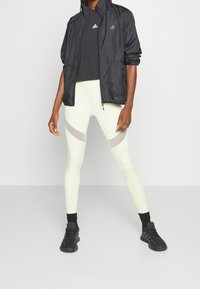 Chaqueta ligera negra, camisa negra y leggings verde claro con detalles en gris. Zapatillas deportivas negras con suelas texturizadas.