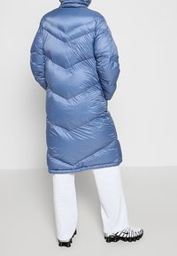 Personne portant un long manteau d'hiver rembourré bleu avec un matelassage en chevron, un pantalon blanc et des chaussures de sport noir et blanc, se tenant de dos.