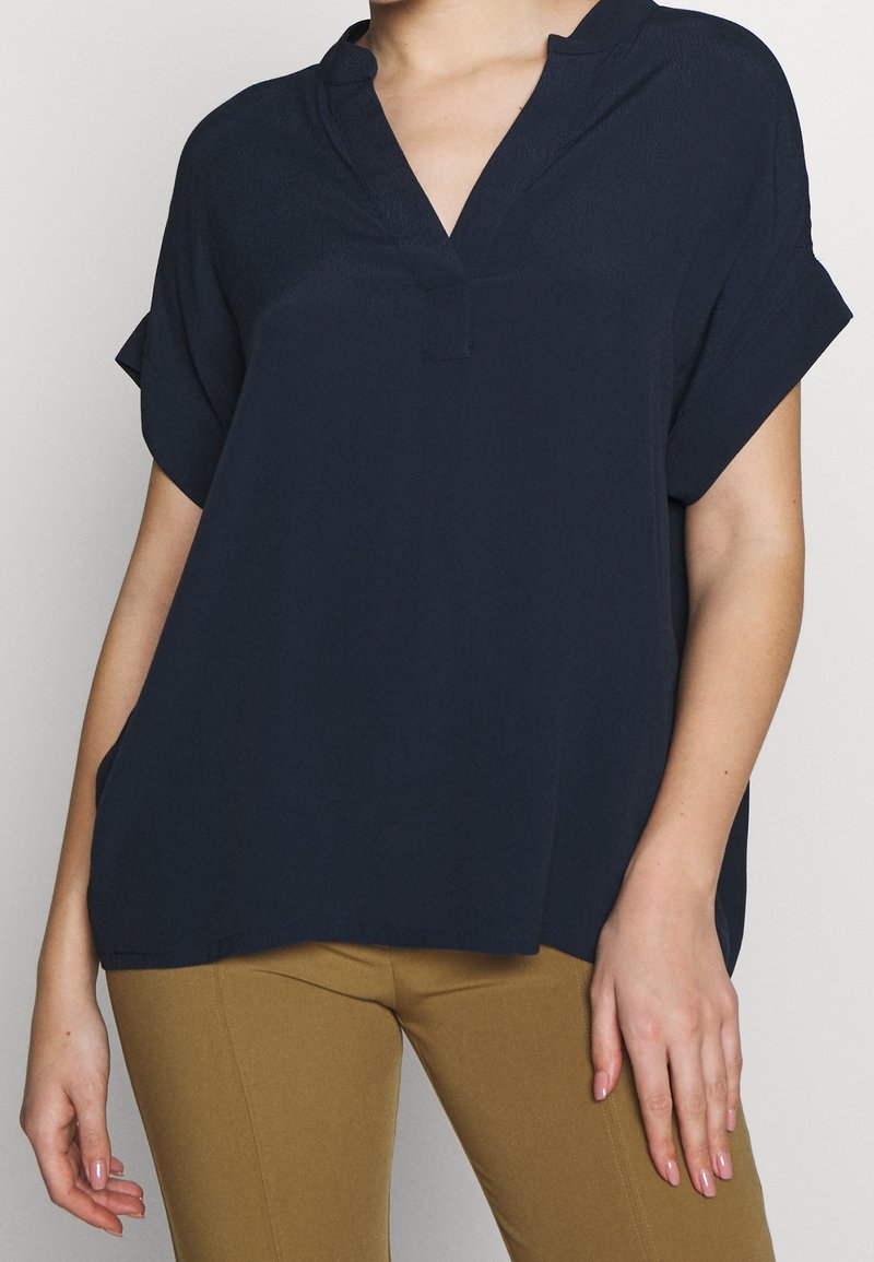 Donna che indossa una blusa nera scura a V con maniche corte e dei pantaloni aderenti color cammello, su uno sfondo semplice.