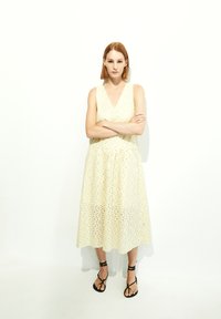 Ärmelloses knielanges Kleid mit V-Ausschnitt in hellem Gelb mit Lochstickerei. Model trägt schwarze Riemchensandalen, die Arme verschränkt.