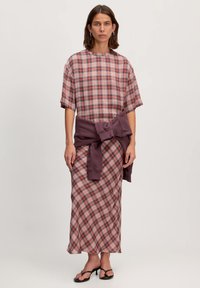 Geruiten jurk in roze, rood en wit, met korte mouwen en een hoge halslijn. Een paarse Sweatshirt is om de taille geknoopt. Sandalen met dunne banden.