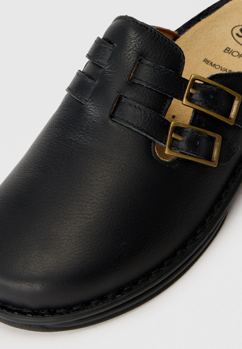 Chaussure noire en cuir à enfiler avec deux boucles en bronze et une finition lisse et texturée. Caractérisée par une semelle intérieure rembourrée et des coutures durables.