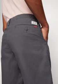 Personne portant un pantalon gris Levi's avec une poche arrière boutonnée et une main dans la poche latérale, montrant la ceinture et les étiquettes de la marque.