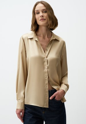 Camicia in satin beige con chiusura a bottoni, maniche lunghe e colletto classico. Texture morbida, design minimalista, abbinata a jeans scuri.
