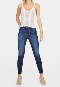 Top de punto blanco sin mangas con escote en V, combinado con pantalones ajustados azul oscuro con detalles desgastados, y sandalias claras con un tacón grueso.