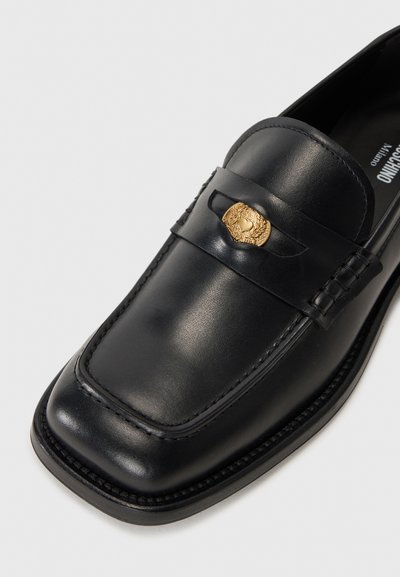 Zwarte leren loafer met een vierkante neus, decoratieve band en een gouden embleem op de wreef. Gladde textuur met contrasterende stiksels.