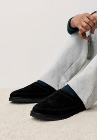 Chaussons en daim noir avec semelle plate et coutures apparentes, portés avec un pantalon de survêtement gris et des chaussettes bleu foncé sur un tapis beige texturé.