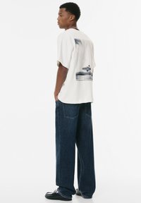 Afslappet-fit creme t-shirt med et blåt grafik på ryggen, parret med løse, mørkeblå jeans og sorte slip-on sko.