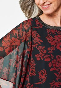 Top nero trasparente con motivo floreale rosso, maniche larghe e scollatura decorativa con perline. Tessuto leggero e design fluido.
