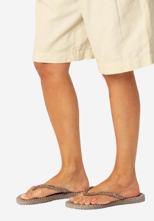 Piernas con pantalones cortos beige que llevan chanclas marrones con suelas tejidas y tiras con cuentas sobre un fondo liso y claro.