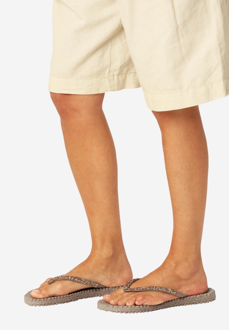 Piernas con pantalones cortos beige que llevan chanclas marrones con suelas tejidas y tiras con cuentas sobre un fondo liso y claro.