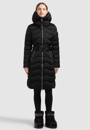 Vrouw die een lange zwarte doorgestikte jas draagt met een hoge kraag, riem, ritszakken, zwarte legging en zwarte winterlaarzen staand tegen een effen achtergrond.