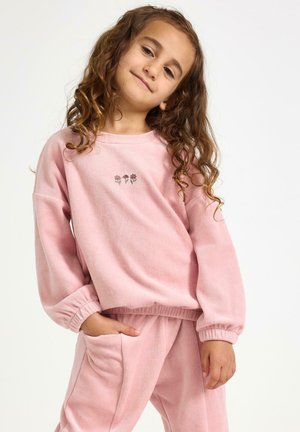 Jeune fille aux longs cheveux bouclés portant un top à manches longues rose et un pantalon assorti, avec une main dans la poche, souriant légèrement.
