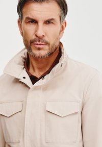 Jasno beżowa kurtka z wysokim kołnierzem, dwoma kieszeniami na klapkę i akcentami w postaci guzików. Wykonana z gładkiego materiału o sztywnym fasonie.