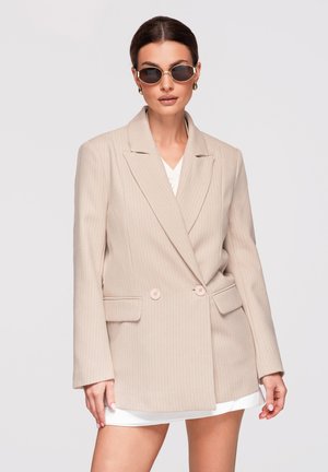 Kvinde med mørkt hår iført beige blazer med nålestriber, hvid top og mørke solbriller, stående mod en ensfarvet lys baggrund.