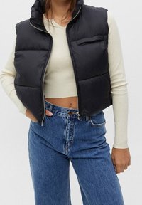 Schwarze Daunenweste mit hohem Kragen, Reißverschluss vorne und einer Brusttasche; getragen über einem cremefarbenen Langarmoberteil und blauen hochgeschnittenen Jeans.