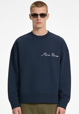 Jonge man met rechthoekige donkere zonnebril en een marineblauwe trui met de tekst "Mira Paris" op de borst, staand voor een witte achtergrond.