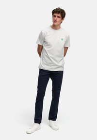 Grå bomull t-skjorte med rund hals og grønt logo, sammen med mørk marineblå bukser og hvite sneakers; modellen står med hendene bak ryggen.