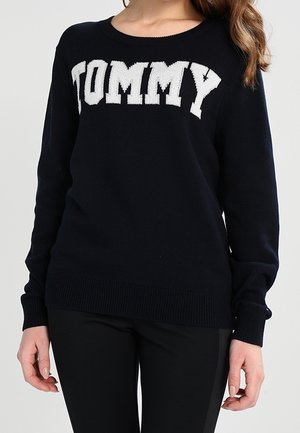 Mujer con un suéter negro con el texto blanco "TOMMY" en el pecho y pantalones negros, de pie frente a un fondo liso.