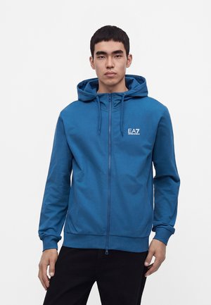 Jeune homme portant un sweat à capuche bleu zippé avec le logo EA7 sur la poitrine, debout devant un fond clair uni.