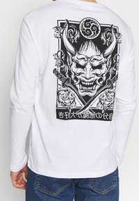 Weißes Langarmhemd mit schwarzer Grafik einer gehörnten Dämonenmaske, gekreuzten Schwertern, floralen Designs und stilisierten asiatischen Zeichen auf der Rückseite.
