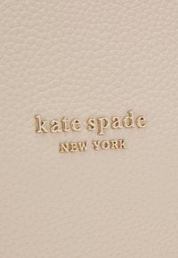 Jasnobeżowa teksturowana skóra z wytłoczonym złotym napisem „kate spade NEW YORK.” Zbliżenie na detal marki na gładkiej powierzchni.