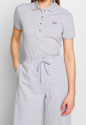 Kobieta w jasnoszarej, krótko rękawowej koszulce polo i pasujących dresowych spodniach ze sznurkiem, stojąca z jedną ręką w kieszeni na jednolitym tle.