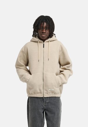 Personne aux dreadlocks mi-longs portant un sweat à capuche beige zippé et un jean foncé, debout les mains dans les poches devant un fond uni.