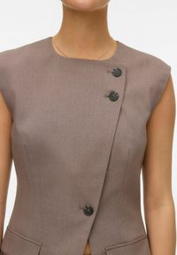 Blazer sans manches marron, présentant un design diagonal, deux boutons noirs, et des poches avant. Le tissu a une texture lisse et mate.