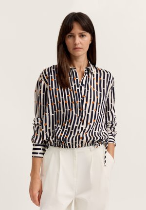 Frau mit glattem braunem Haar trägt eine langärmelige, gemusterte Bluse mit schwarzen und orangen Streifen, kombiniert mit hoch taillierter weißer Hose.