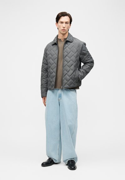 Νέος άντρας στέκεται φορώντας γκρι πομπε μπουφάν, ελιά πουλόβερ, φαρδιά ανοιχτό μπλε τζιν και μαύρα παπούτσια πάνω σε απλό λευκό φόντο.