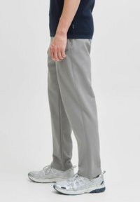 Uomo che indossa pantaloni grigi chiari e scarpe da ginnastica argentate, in piedi contro uno sfondo bianco uniforme, vista di lato.