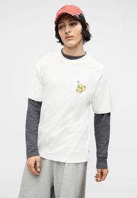 Νεαρός που φοράει κόκκινο καπέλο μπέιζμπολ, άσπρο μπλουζάκι με μικρό γραφικό Pikachu, γκρι μακρυμάνικο πουκάμισο από μέσα και γκρι παντελόνι, όρθιος.