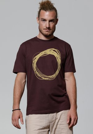 T-shirt en coton bordeaux avec un design graphique circulaire doré. Manches courtes, col rond, coupe décontractée. Texture douce avec un aspect informel.