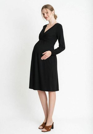 Zwangere vrouw in een zwarte jurk met lange mouwen en bruine sandalen met hak, die zachtjes haar buik vasthoudt en glimlacht, staand tegen een witte achtergrond.