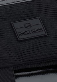 Musta teksturoitu kangas, jossa on sileä ja kiiltävä osio; siinä on musta kumitag, jossa lukee kohokuvioituina kirjaimina "JOHNNY URBAN".