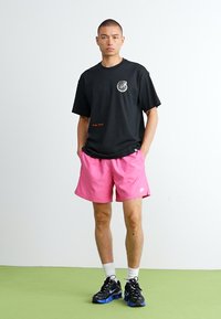 Sort t-shirt med rund hals og et logo-patch, parret med pink shorts. Modellen har sorte og blå sneakers på en grøn overflade.