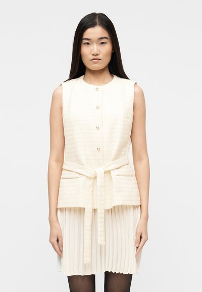 Femme aux longs cheveux noirs portant un gilet crème texturé sans manches boutonné avec une ceinture nouée, sur une jupe plissée crème.