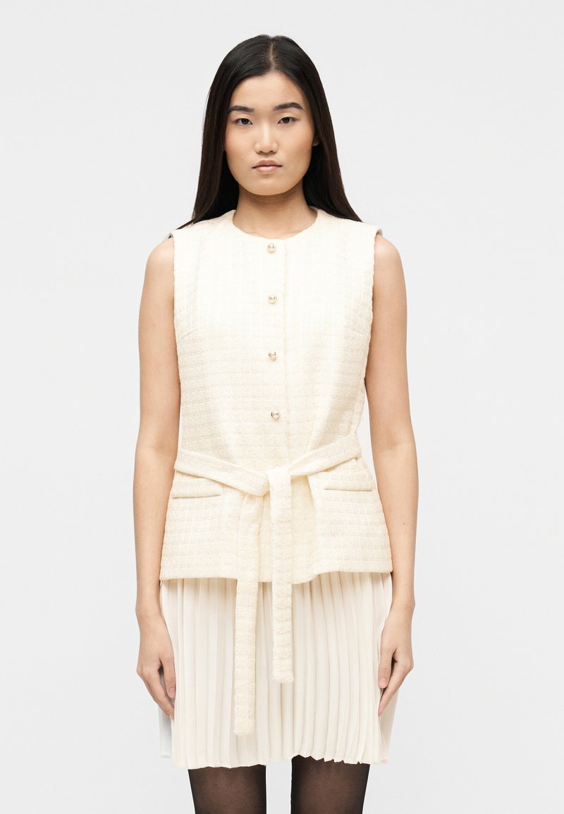 Γυναίκα με μακριά σκούρα μαλλιά πο υ φοράει αμάνικο εκρού υφασμάτινο γιλέκο με κουμπιά και ζώνη δεμένη πάνω από πλισέ εκρού φούστα.