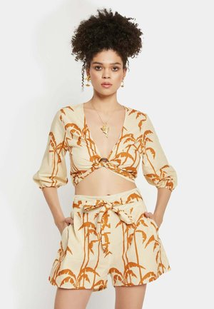 Cremfarbene Kleidung mit Palmenmuster in rostorange. Das verkürzte Oberteil hat einen tiefen V-Ausschnitt und ein geknotetes Design; die Shorts sind hoch tailliert und mit einer Schleife versehen.
