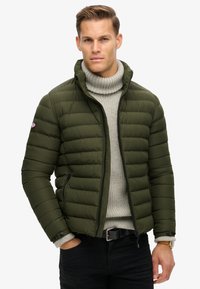 Oliven grøn pufferjakke med vandrette sømme, stående krave og lynlåslommer, båret over en lys grå ribbestrikket sweater.
