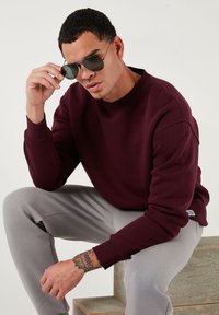 Bordeaux sweatshirt met een ronde hals en geribbelde manchetten, gecombineerd met lichtgrijze sweatpants. Metalen zonnebril accentueert de look.
