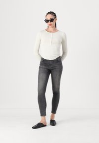 Bijela teksturirana majica s dugim rukavima i detaljem gumba, u kombinaciji s crnim skinny trapericama i crnim slip-on cipelama; model nosi kutne crne sunčane naočale.