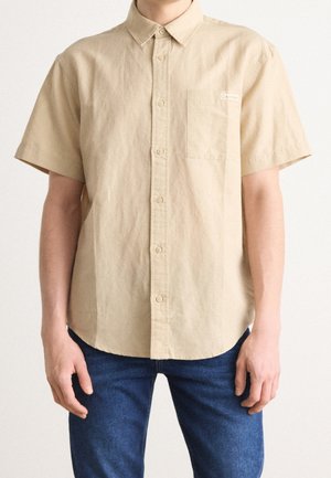 Camicia beige a maniche corte con bottoni e taschino anteriore, indossata da persona con jeans blu, in piedi davanti a uno sfondo semplice.