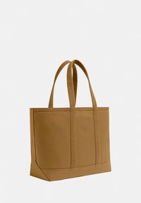 Sac cabas en toile marron avec deux longues anses et coins inférieurs renforcés sur un fond blanc uni.