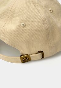 Gorra beige de algodón con un acabado texturizado. Presenta una hebilla de latón y un acento de logo en la correa trasera.