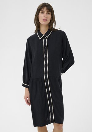 Kobieta ubrana w czarną sukienkę zapinaną na guziki z beżowym zygzakowatym wykończeniem na kołnierzu, przodzie i mankietach, stojąca na tle jednolitej białej ściany.