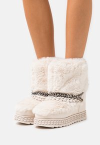 Botas crema de piel sintética con una suela gruesa y texturizada, decoradas con un detalle de cadena sobre el empeine y un amplio puño mullido en la parte superior.