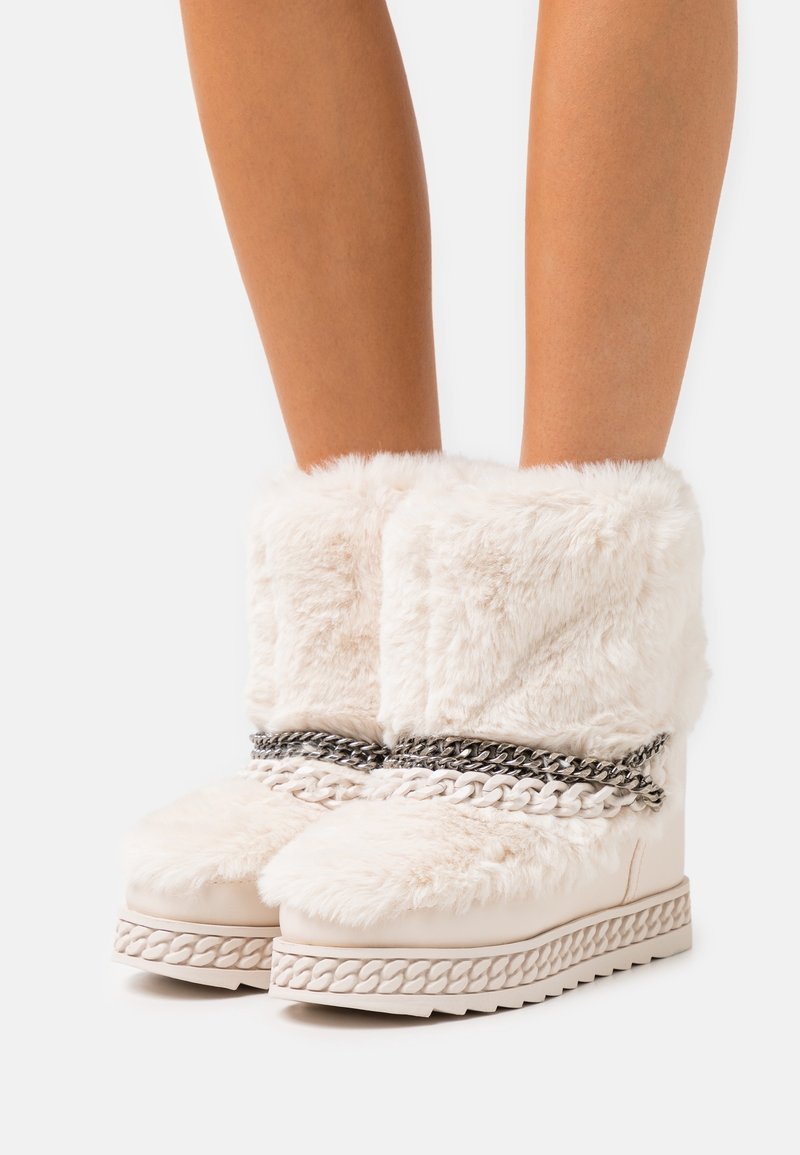 Botas crema de piel sintética con una suela gruesa y texturizada, decoradas con un detalle de cadena sobre el empeine y un amplio puño mullido en la parte superior.
